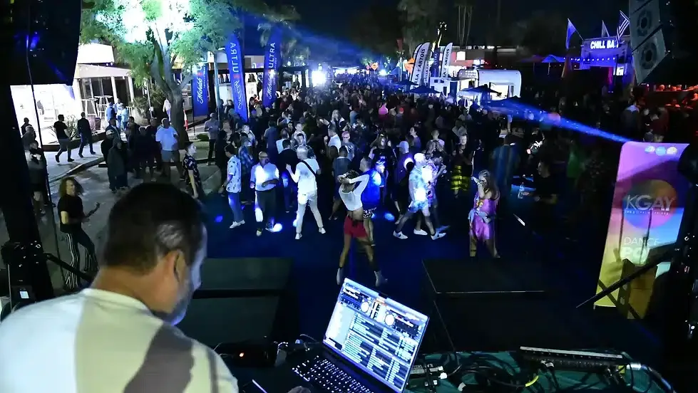 A DJ is seen from behind looking out over a lively nighttime outdoor party scene with vibrant lighting and a crowd of people dancing and socializing.