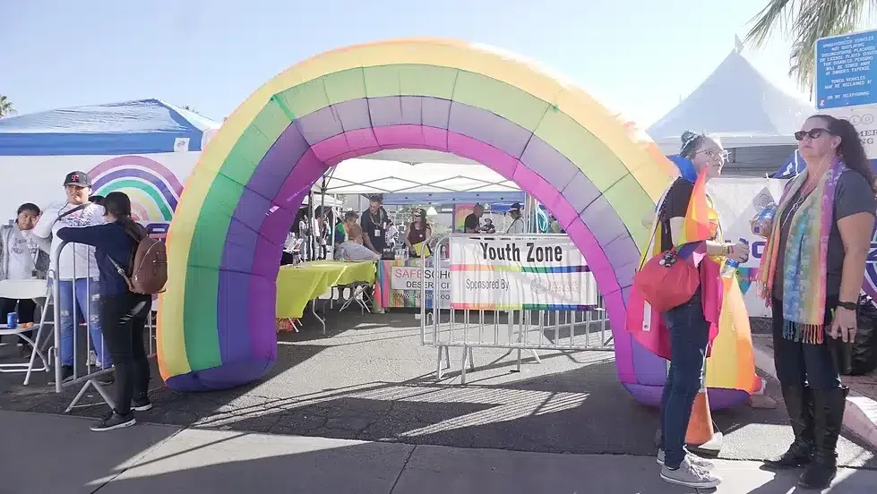 An outdoor event with people standing near an inflatable rainbow archway entrance labeled "Youth Zone." The attendees are casually dressed, some wearing rainbow motifs, with vendor booths and blue skies in the background.