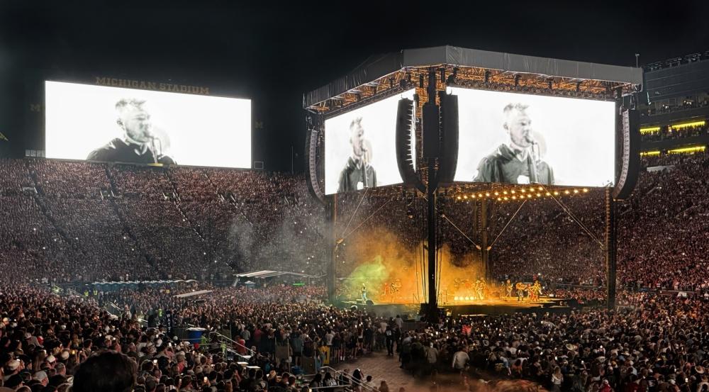 Night-time concert. Band performs in the center of a stadium with a jumbotron above the band with two screens streaming the performers. Another screen spans across stadium seating in the background. The audience holds up phones and lights to illuminate the stadium.