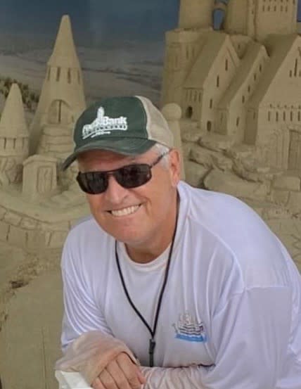 A middle-aged man in a baseball cap, a long-sleeve white shirt, and dark sunglasses leans over in front of a sand sculpture and smiles at the camera.