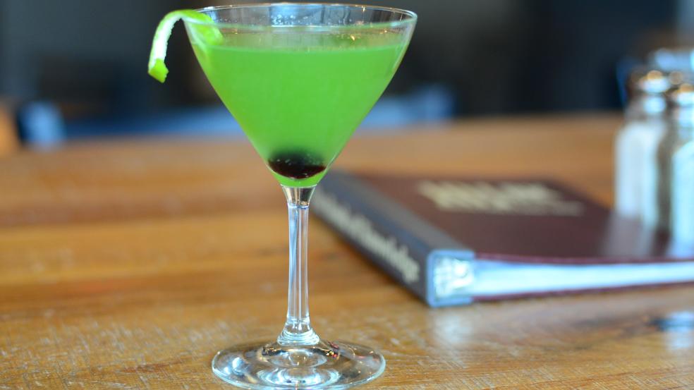 Bright green cocktail in a martini glass with cherry at borrom and lime twist garnish.
