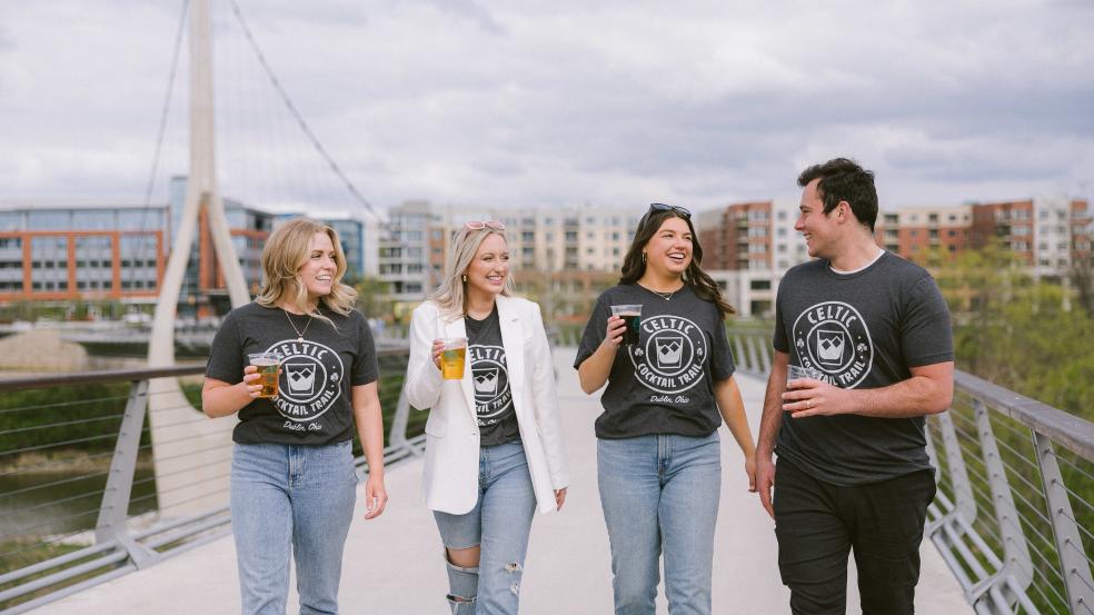 A group of friends walking across the Dublin Link pedestrian bridge with DORA cups in Celtic Cocktail Trail t-shirts