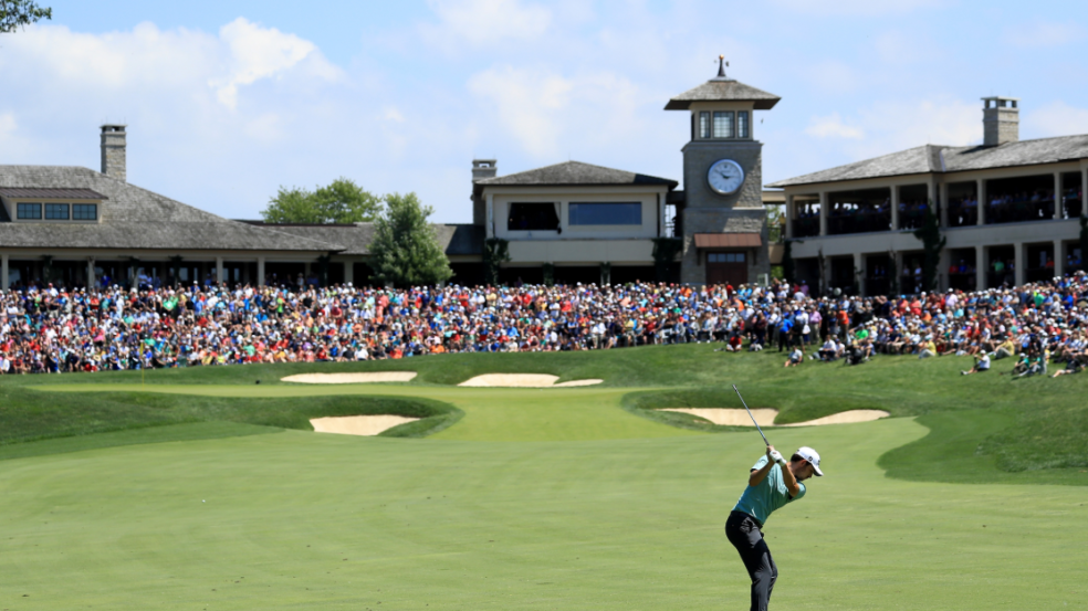 Golfer swining club at golf ball towards the 18th green that is surrounded by a crowd at the Memorial Touranemnt.