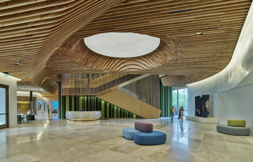 A grand, modern lobby featuring a wavy slatted wood ceiling with a circular cutout, a large wooden staircase, and a curved reception desk with a green textured front. Light-colored stone floors, colorful ottomans, and large windows with people and trees complete the scene.