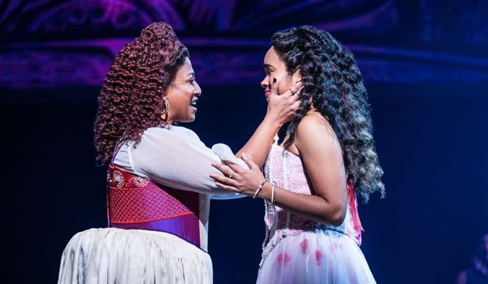 Two women stand on stage, the one on the left has her arm extended holding the face of the one on the right, smiling while she sings. The woman on the right holds the arms of the one on the left.