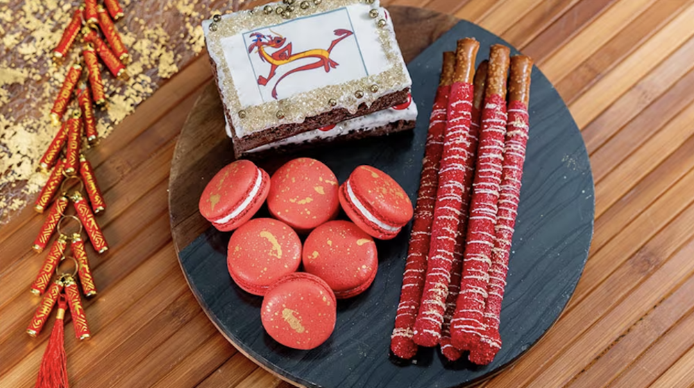 Image of three desserts on a black plate. The desserts look to be red cookies, red pretzel sticks dipped in chocolate, and a piece of cake. The plate is placed upon a wooden background.