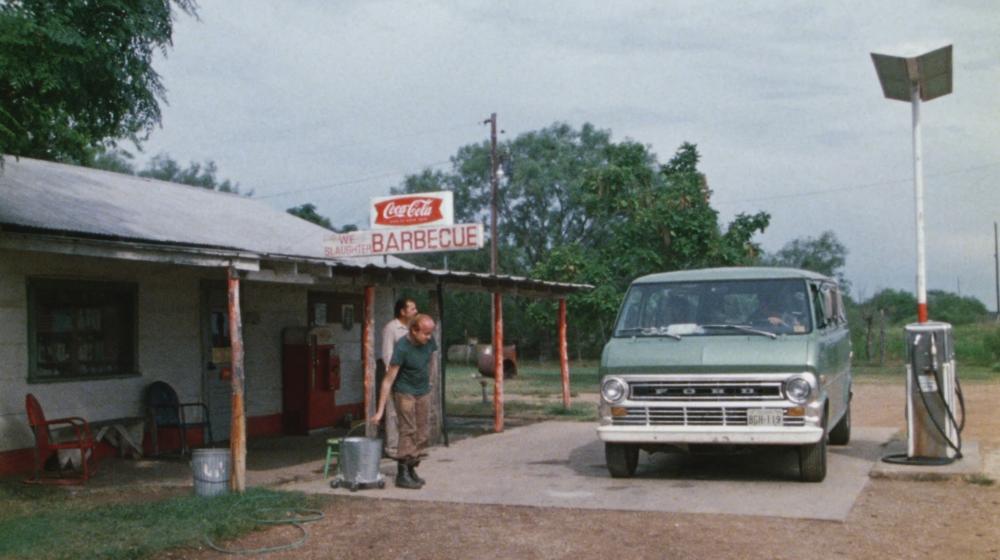 Texas Chainsaw Massacre screengrab, showing a green van parked in front of a gas station with a coca cola and Barbecue sign. Two men walk towards the van