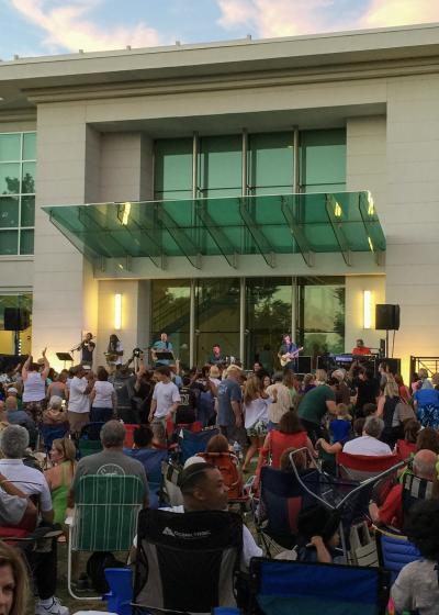 A crowd gathers on the lawn in Big Spring Park for Concerts in the Park, relaxing in lawn chairs and dancing while a live band performs on an outdoor stage in front of a modern building at sunset.