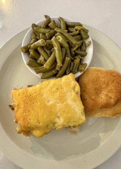 A plate of homestyle comfort food from Main Street Café features a baked chicken casserole topped with melted cheese, a side of seasoned green beans, and a soft dinner roll. The meal is served on a white plate atop a white tablecloth.