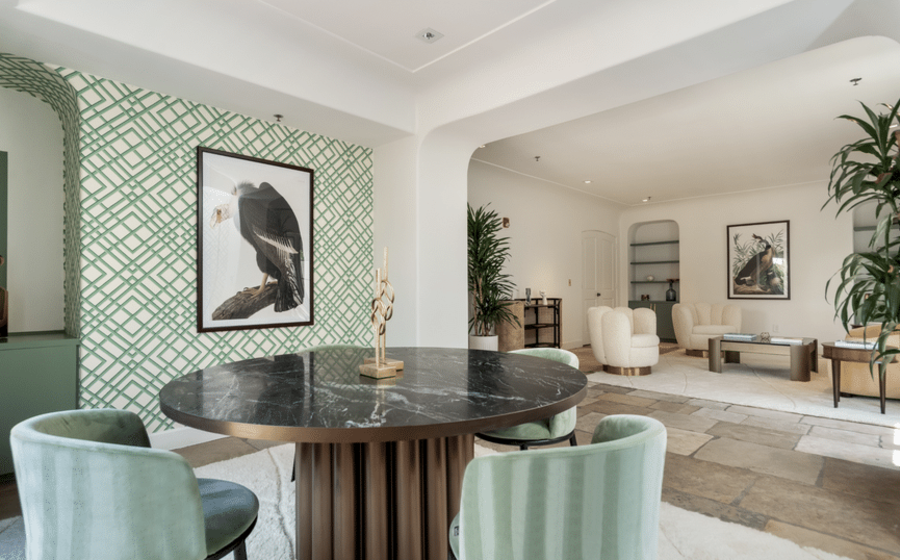 A modern, spacious living room with a black marble round table and green velvet chairs in the foreground. The walls are decorated with geometric-patterned wallpaper and framed bird photographs. The background shows a seating area with cream-colored armchairs, plants, and tasteful decor.