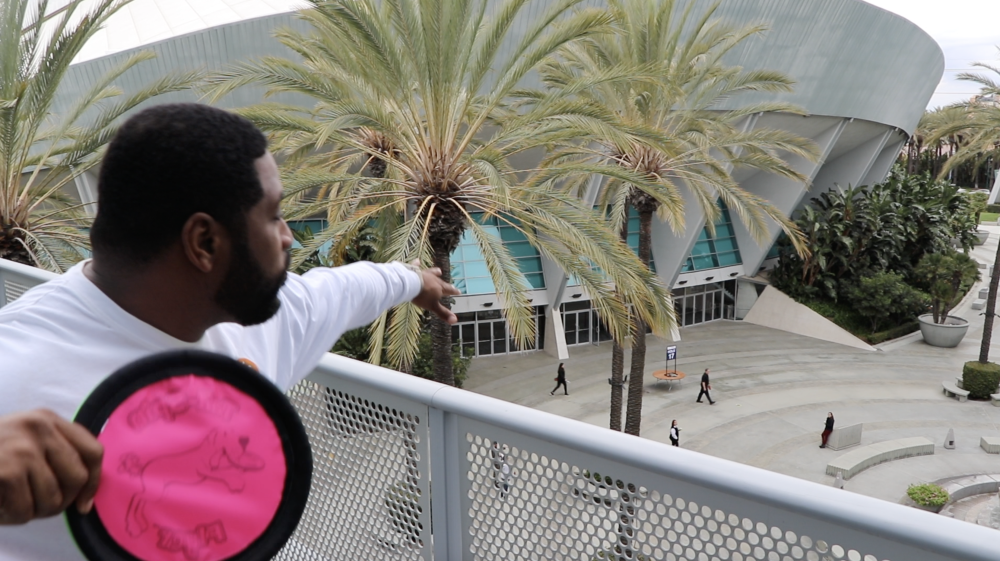 Image of a pink frisbee being thrown off a balcony by a gentleman in a white shirt. The gentleman is participating a disc golf tournament.