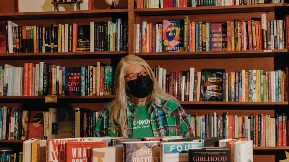 A woman wearing a black face mask stands in front of wooden bookcases lined with books at BookWoman