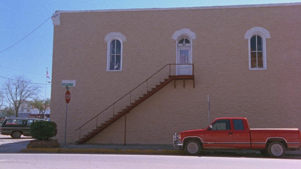 Waiting for Guffman screengrab showing a red truck parked on the side of a two story building. The building has a metal staircase leading up to a white door Corky's Apartment
