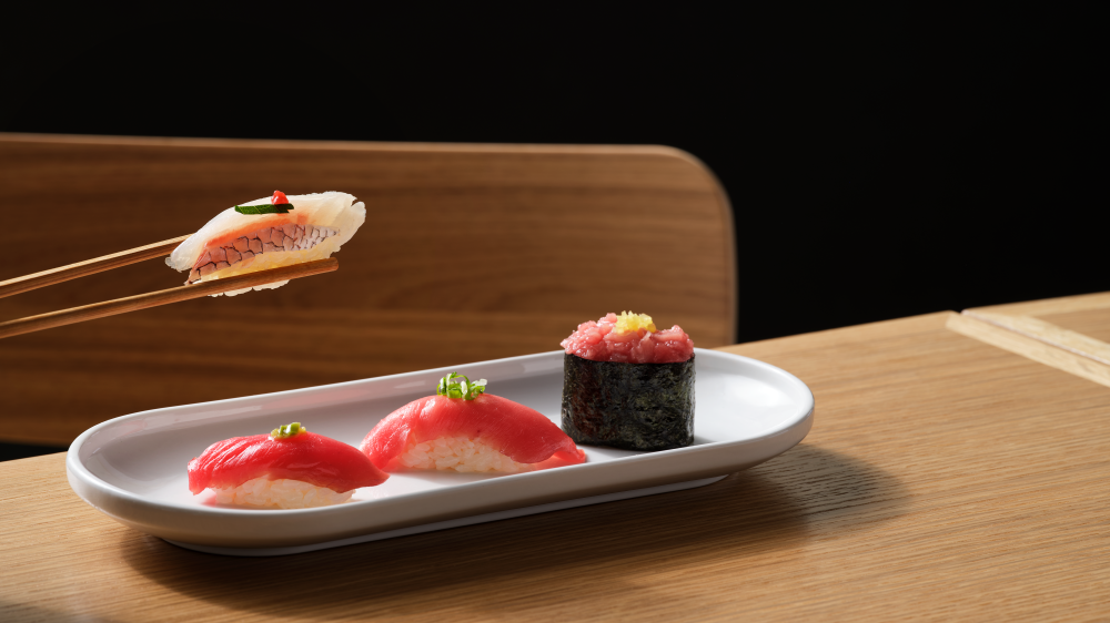 Close-up image of two chopsticks holding a sushi roll with three other pieces of sushi sitting on a plate.