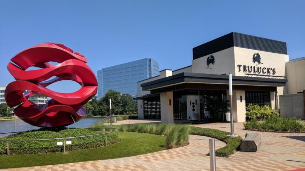Truluck's is an angular building with a black-and-cream color scheme. To the left of the entrance is a large red sculpture that looks like ocean waves wrapped into a ball. Behind it is Lake Woodlands and a bright blue sky.