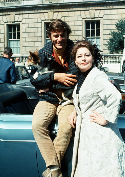 Roddy McDowall holding Ava's corgi standing next to Ava Gardner in front of a car on set of Tam Lin in London.