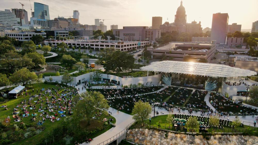 Aerial view of Moody Amphitheater in evening sun. There is a crowd on the open lawn and in seats near the pavilion stage. The State Capitol building is visible behind the amphitheater
