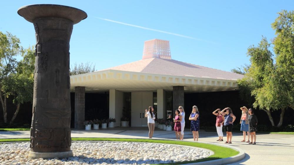 A group of people standing outside a modern building with a distinctive roof and an ancient-styled column in the foreground.