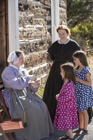 Two women in pioneer dresses talk to two girls outside a log cabin at Golden History Museum & Park