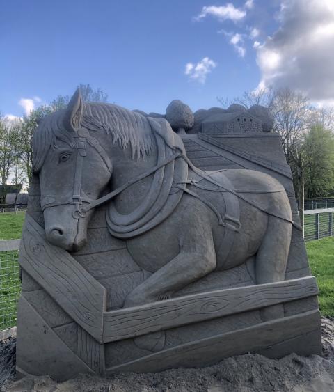 Sand sculpture positioned in a green space that shows a working horse coming 3-D out of the sculpture. There is also a fence and a barn visible in the sand behind the horse.
