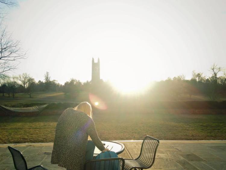 A young woman sits outside at a table with chairs and the Princeton University tower in the background
