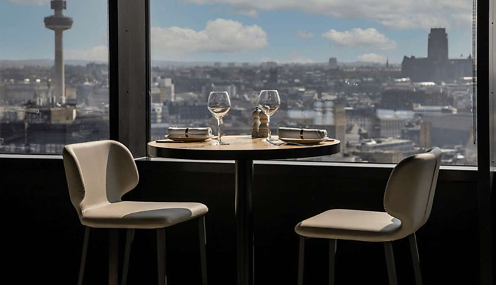 2 high chairs and a table with 2 wine glasses on top of it, positioned next to a big window where you can see Liverpool's skyline.