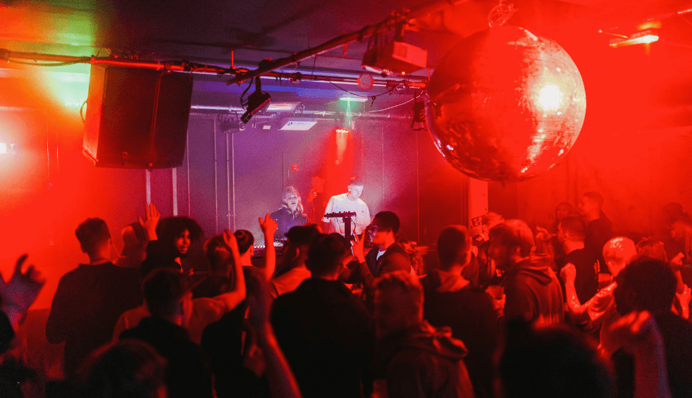 Inside a club with a large disco ball, red lights and a crowd dancing.