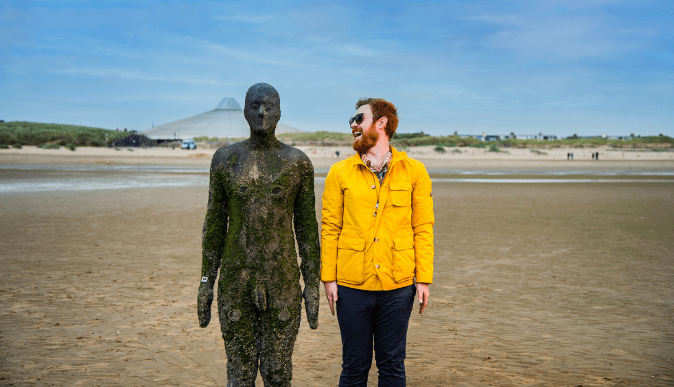 A man in a yellow raincoat stands next to a cast-iron statue on Crosby beach