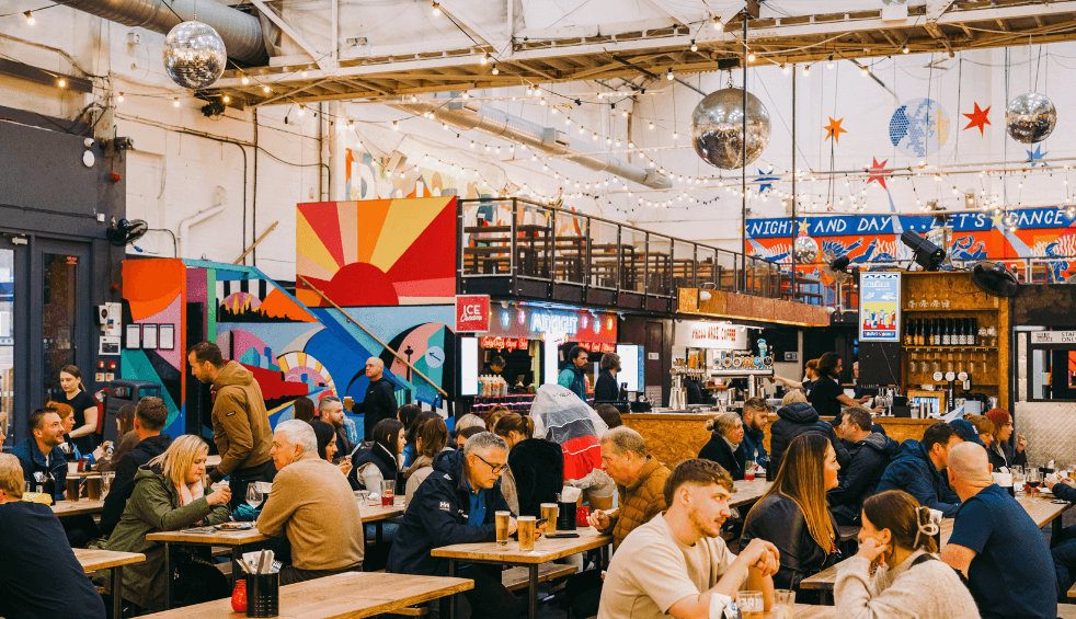 People sitting at benches in a large warehouse that has been transformed into a vibrant food marker. There are disco balls hanging from the ceiling.