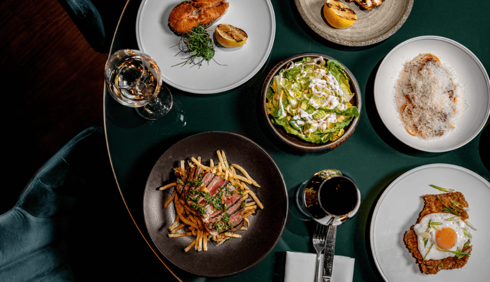 A top down view of a table with plates of food on including steak and chips, salad and a fried egg.