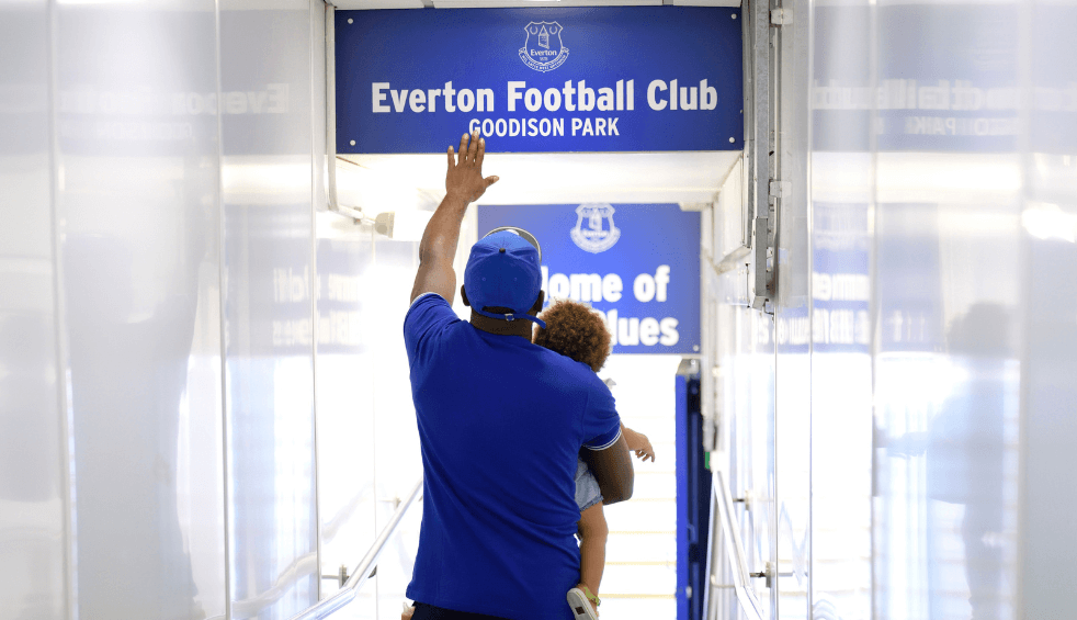 A person carrying a small child walking down the tunnel at Everton Stadium touching an Everton sign about the door.