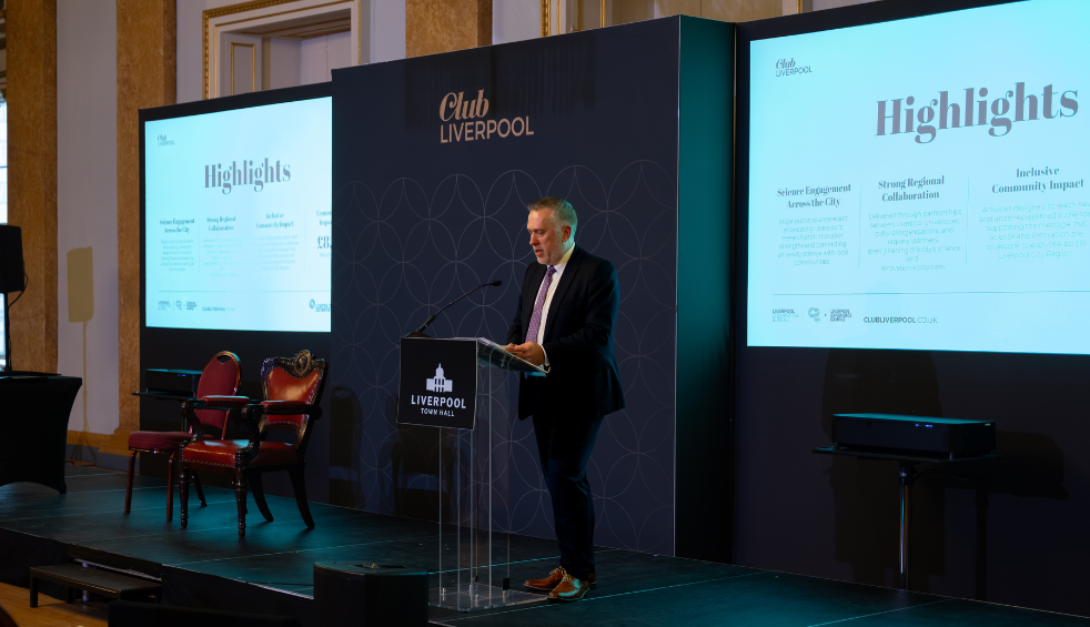 Person standing at a lectern on a stage at Liverpool Town Hall, speaking in front of ‘Club Liverpool’ branded panels and two presentation screens titled ‘Highlights’, with chairs positioned to one side of the stage.