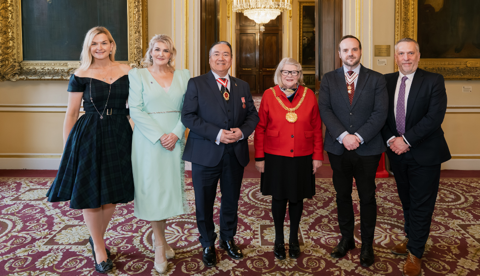 Group of people standing in a formal reception room with ornate décor and framed paintings, dressed in formal attire, with some individuals wearing ceremonial chains and medals.