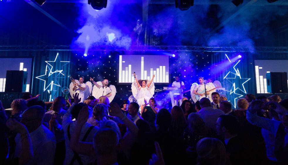 Wide view of a crowded indoor concert with a live band performing on a stage lit in blue and purple, featuring illuminated star-shaped light frames, smoke effects, and a central LED screen displaying animated equaliser graphics, while audience members raise their hands in the foreground.