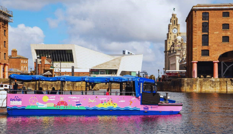 A pink and blue boat that looks like a bus floating on a body of water at docks in Liverpool. The Royal Liver Building is in the background.