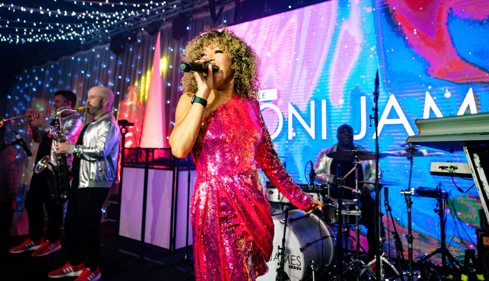 Live band performing on stage under string lights and colourful LED screens, with a lead singer in a bright pink sequined outfit singing into a microphone at centre, musicians playing brass, drums, and keyboards behind, and large screen graphics reading “Toni James” in the background.