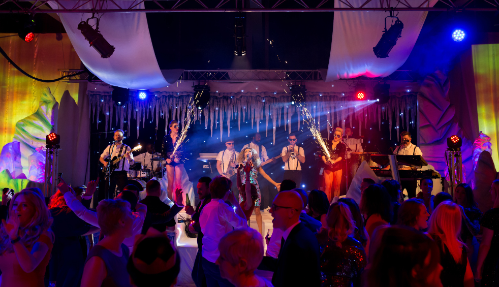 Wide view of a live band performing on a decorated indoor stage with colourful lighting, hanging fabric panels, and icicle-style set pieces. Performers stand across the stage playing instruments while sparkler-style pyrotechnics fire upward, and a large audience fills the foreground, watching and raising hands.