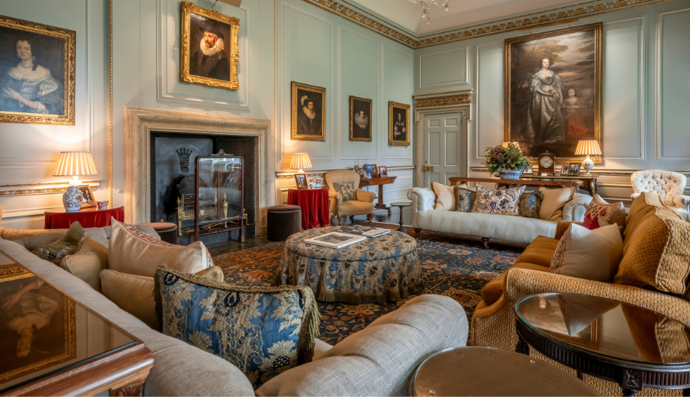 Elegant historic interior showing a grand sitting room with pale blue panelled walls, ornate mouldings, and framed oil portraits. Plush sofas and armchairs surround a central upholstered ottoman on a patterned rug, with side tables, lamps, and a fireplace creating a formal, stately atmosphere.
