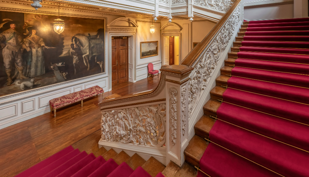 Ornate historic interior featuring a sweeping staircase with carved stone balustrades and deep red carpet runners, polished wooden floors, and large classical paintings displayed along the walls, lit by warm hanging lamps and ceiling lights.