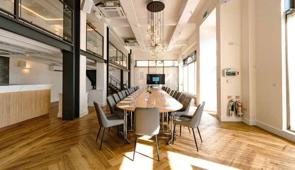 Bright, open-plan meeting and event space featuring a long boardroom table, wooden floors, high ceilings with modern pendant lights, large windows letting in natural light, and a presentation screen at the far end.