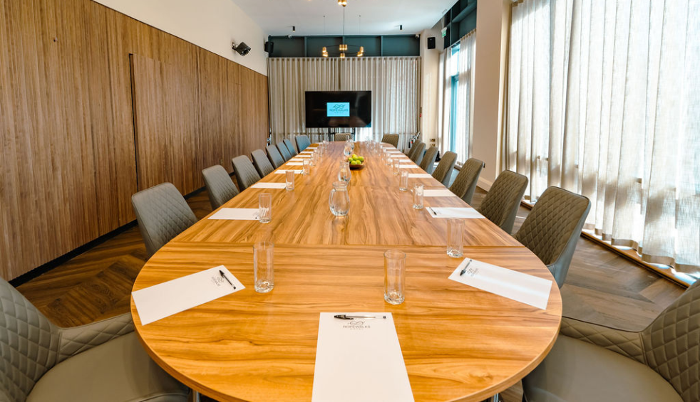 Long wooden conference table prepared for a meeting, laid with branded notepads, pens, water glasses and a central fruit bowl, with padded chairs and wood-panelled walls along one side.