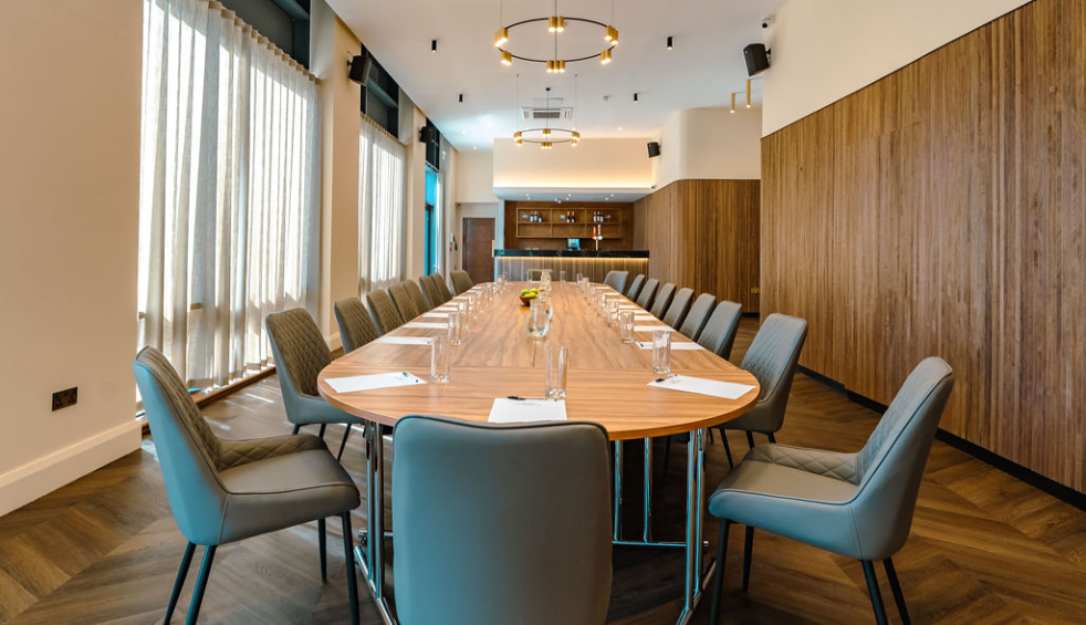 Modern boardroom with a long oval wooden table set with notepads, pens, water glasses and jugs, surrounded by upholstered chairs, with floor-to-ceiling curtains and warm pendant lighting overhead.