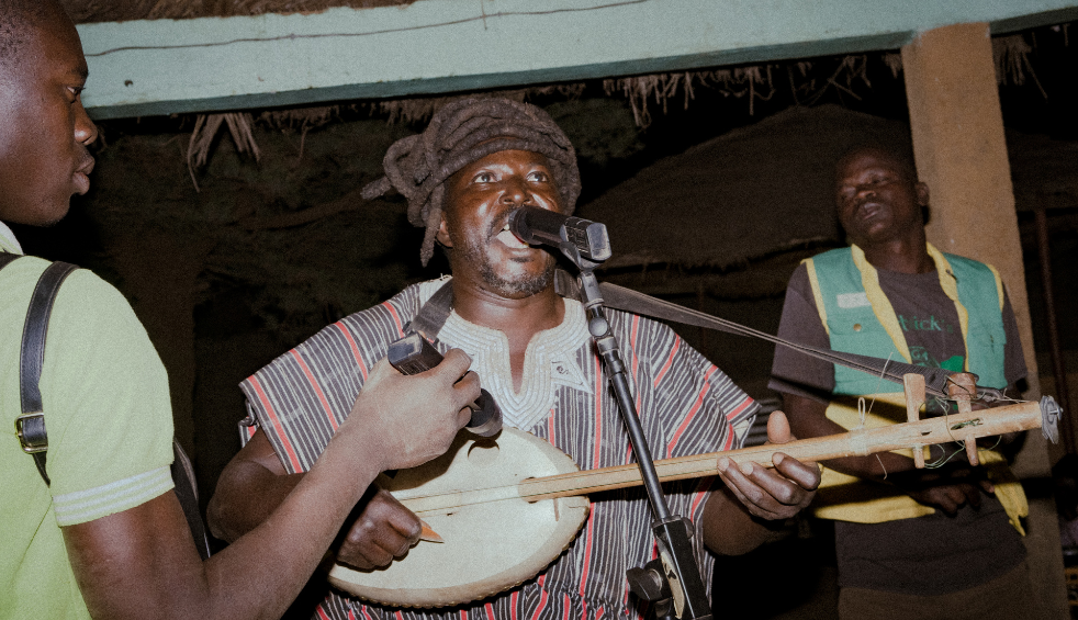 Indoor performance setting showing a musician singing into a microphone while playing a traditional stringed instrument. Two other musicians stand nearby, one holding percussion. The scene is lit with warm, low lighting beneath a thatched roof structure.