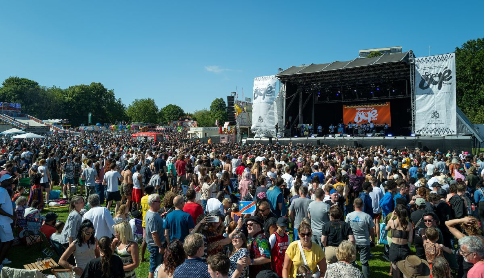 Wide outdoor festival scene with a large crowd standing on grass in front of a main stage. The stage features lighting rigs, banners, and a large central screen, with trees and fairground rides visible in the background under a clear blue sky.