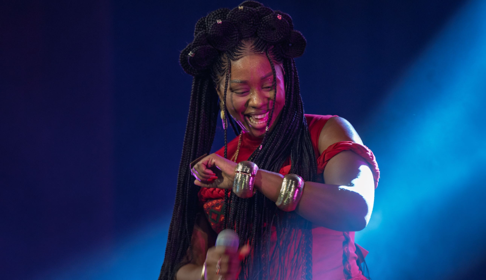 Close-up of a live stage performance showing a performer holding a microphone while wearing a red outfit. Long braided hair is styled up, and metallic wrist cuffs are visible. Blue and purple stage lighting illuminates the background.