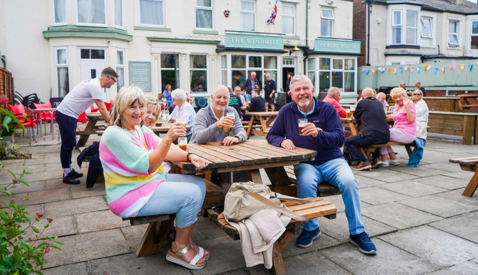 People seated at wooden picnic benches outside a pub with signage reading “The Windmill Public House,” holding drinks and socialising in a courtyard setting with bunting decorations and other guests in the background.