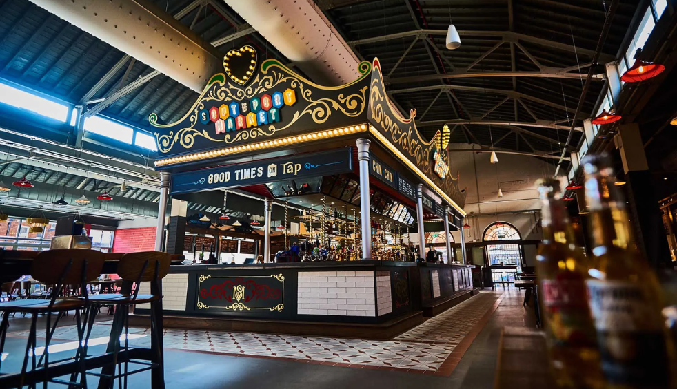 An indoor food hall bar with ornate signage reading “Southport Market” and “Good Times on Tap,” featuring a central bar island, high stools, patterned tiled flooring, and industrial-style ceilings with exposed beams and hanging lights.