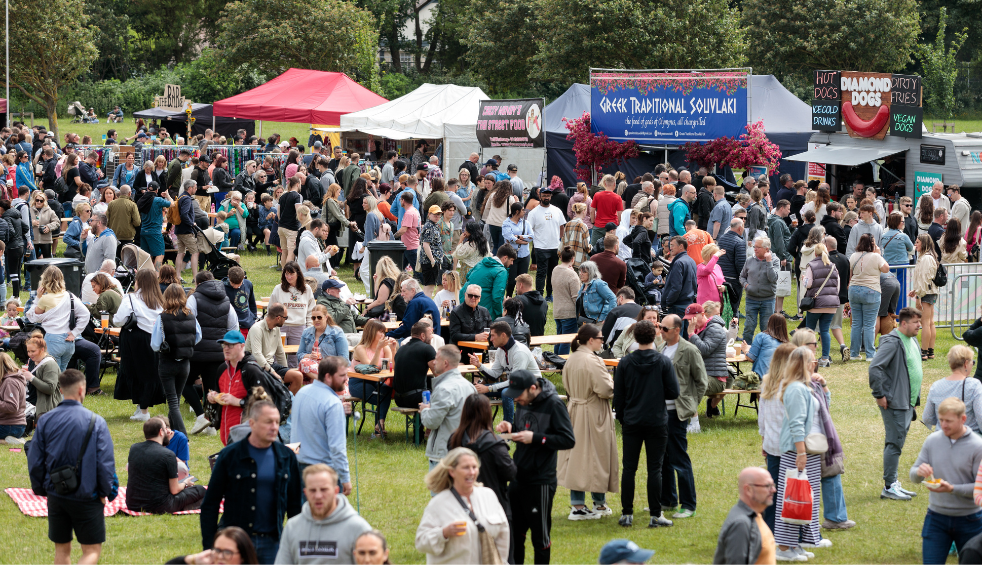A large crowd gathers at the Southport Food and Drink Festival, spread across a grassy park filled with rows of food stalls and colourful tents. Visitors stand, walk, and sit at wooden benches eating and drinking, while vendor signs advertise international cuisine, including Greek dishes and street food. Trees line the background, creating a busy, open-air festival atmosphere on a bright day.