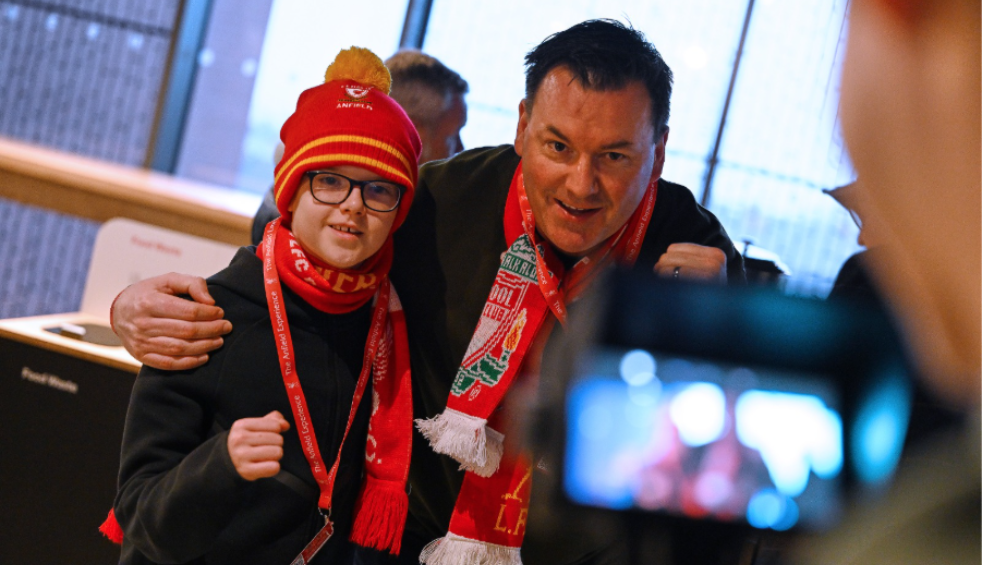 A man and young boy getting their photo taken in Liverpool Football Club stadium with the LFC scarf and hat on.