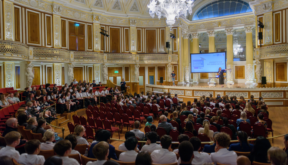 Ornate auditorium with gold detailing and chandeliers, filled with seated attendees facing a stage where a presenter stands beside a large projection screen. Rows of red chairs fill the space, with additional audience members seated along a balcony and side tiers.
